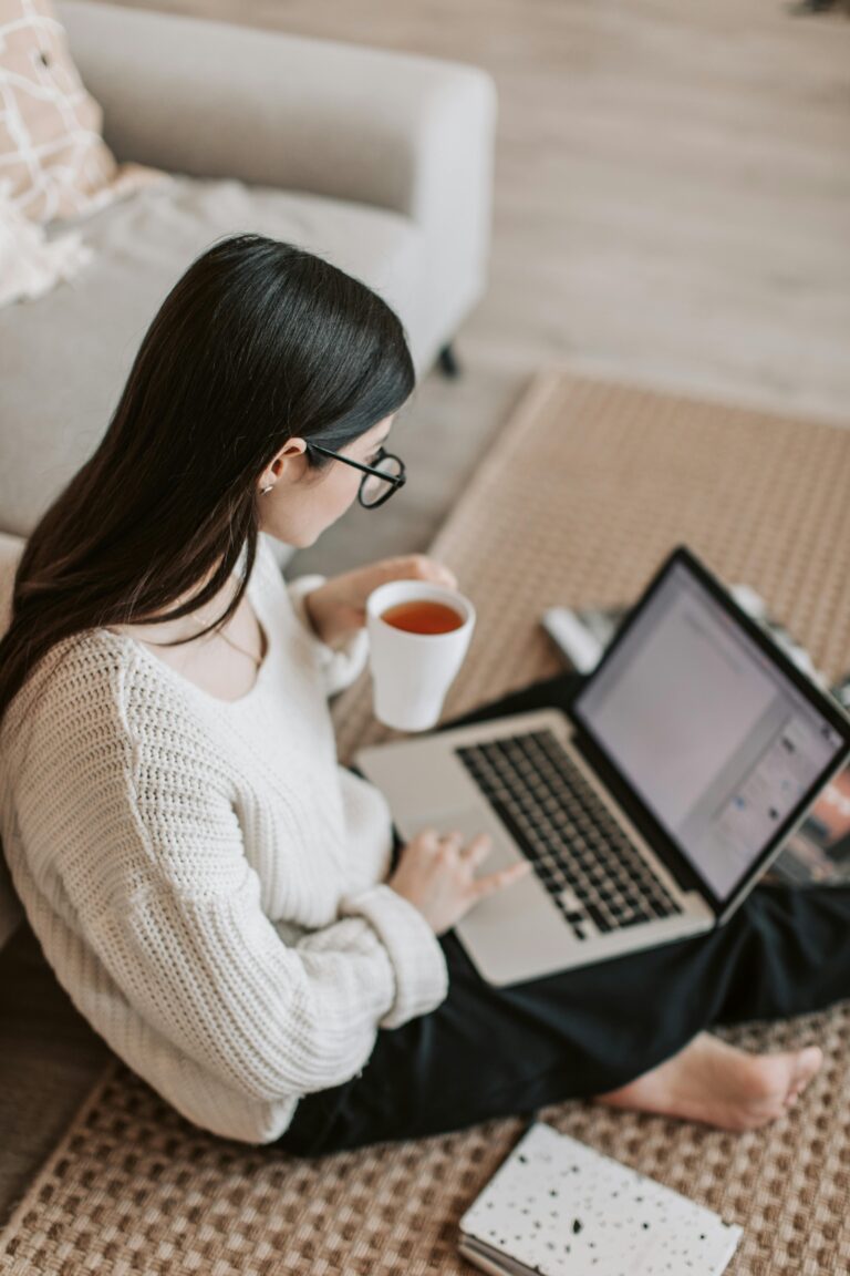 Adult woman in cozy sweater works on laptop while enjoying a hot drink indoors.